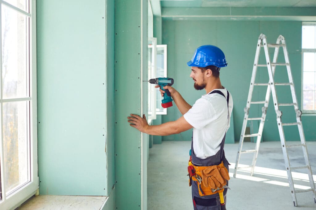 Worker builder installs plasterboard drywall at a construction site office open space. Plasterboard installer.