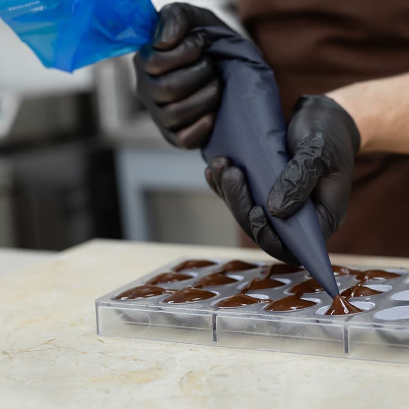 Close-up of the chocolatier's hands pouring chocolate into the molds. The chef's pastry bag pours hot melted chocolate into a silicone mold.
